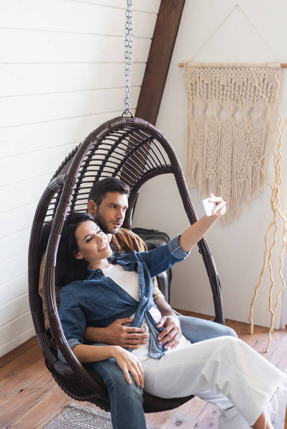 heureux jeune couple assis dans une chaise en rotin suspendu et prendre selfie sur téléphone mobile - Photo, image