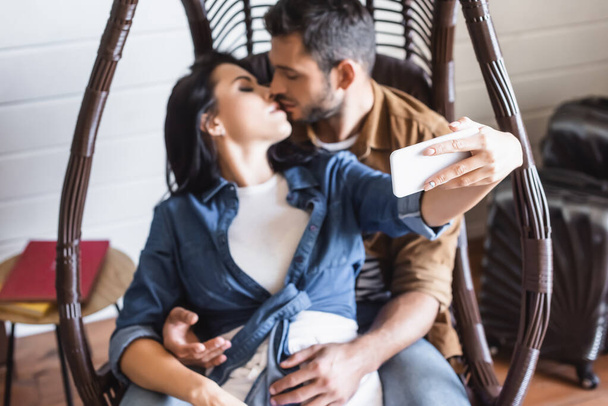 jeune femme prenant selfie sur smartphone tout en embrassant avec petit ami dans une chaise suspendue, fond flou - Photo, image