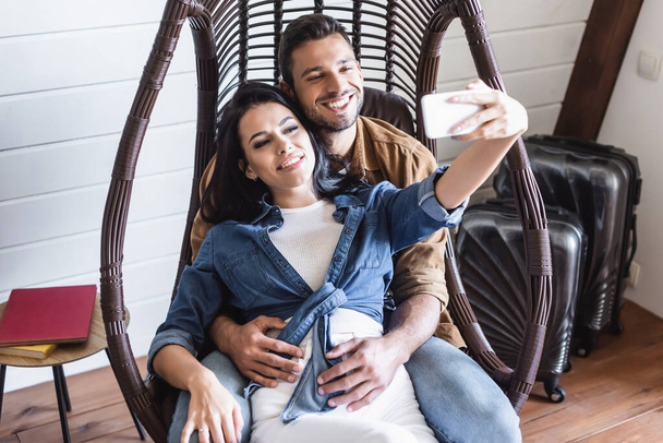 couple joyeux prenant selfie sur smartphone tout en étant assis dans une chaise en rotin suspendu - Photo, image