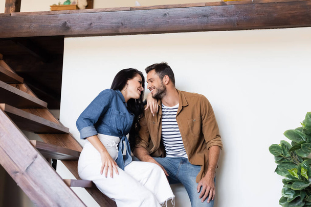 happy couple in stylish casual clothes looking at each other on wooden stairs at home - Φωτογραφία, εικόνα