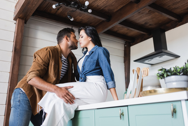 young man touching sensual woman sitting on kitchen counter in stylish casual clothes - Foto, afbeelding