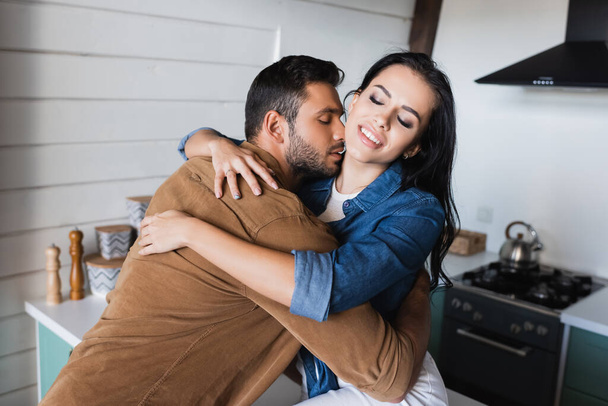 happy brunette woman with closed eyes hugging man kissing her neck in kitchen - Foto, imagen