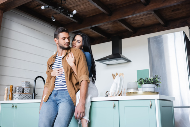 happy young woman sitting on kitchen counter and embracing boyfriend  - Foto, Imagen