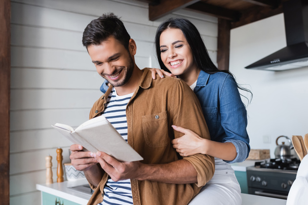 laughing woman hugging arm of cheerful boyfriend reading book in kitchen - Photo, Image