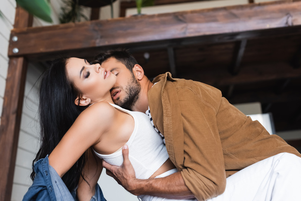junger Mann umarmt und küsst sexy brünette Frau in stilvoller Kleidung zu Hause - Foto, Bild