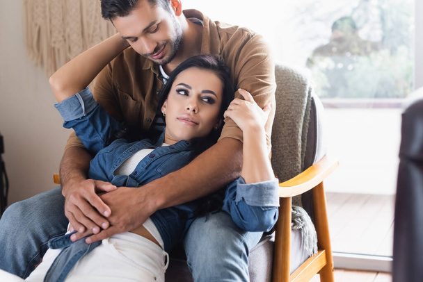 happy man sitting in armchair and embracing girlfriend at home - Photo, Image