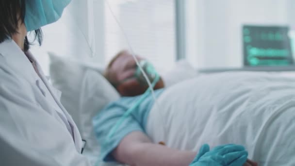 Detailní záběr ženského lékaře v ochranném obličejovém skle, masce a rukavicích sedících na ošetřovně a držících se za ruku mužského pacienta ležícího na ventilátoru během léčby covid-19 - Záběry, video