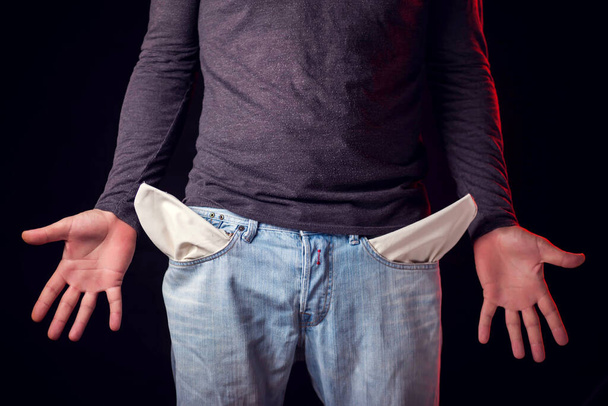 Man toont lege broekzak van zijn jeans voor zwarte achtergrond. Armoede en financiële problemen - Foto, afbeelding