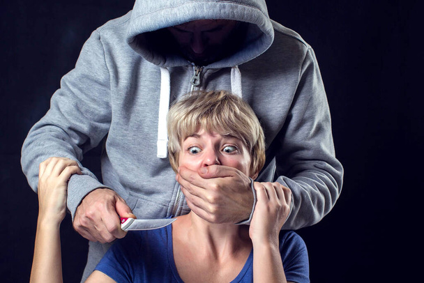 Hombre con suéter con capucha que cubre la boca de una mujer y sostiene un cuchillo. Concepto de violencia o secuestro.  - Foto, imagen