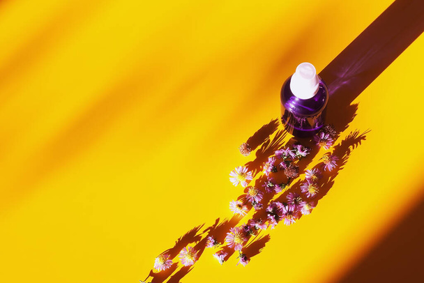 Sarı arka planda çiçekler olan mor sıvı kozmetik şişesi. Düz yat, boşluğu kopyala - Fotoğraf, Görsel