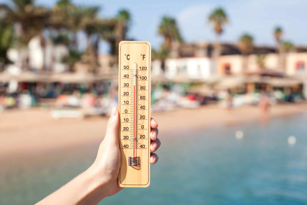 Mano de mujer sostiene termómetro en el fondo de la playa. Concepto de verano, clima y clima - Foto, imagen