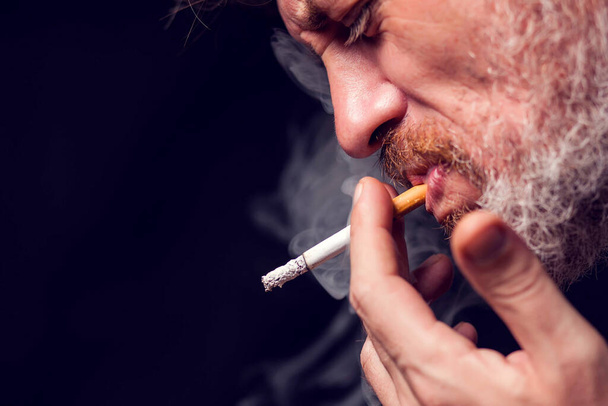 Muotokuva miehestä polttaa savuketta mustalla pohjalla. Tupakkariippuvuuden käsite - Valokuva, kuva