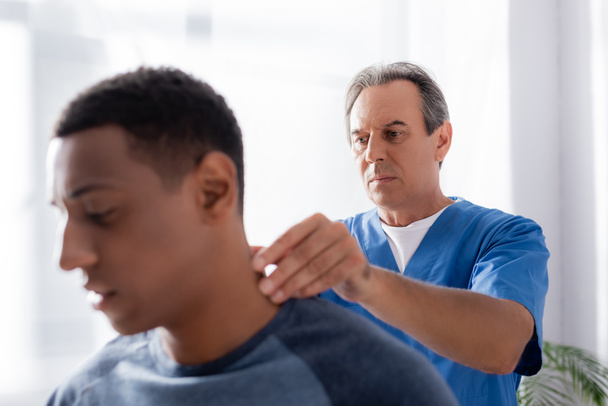 masajista masajeando lesionado cuello de africano americano paciente en borrosa primer plano  - Foto, Imagen