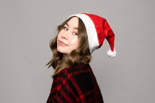 Kırmızı kazaklı ve chritmas şapkalı gülümseyen beyaz kadın portresi gri arka planda doğal makyajlı kameraya bakıyor. Yeni yıl, Noel tatili. Kıvırcık saçlı kız. - Fotoğraf, Görsel