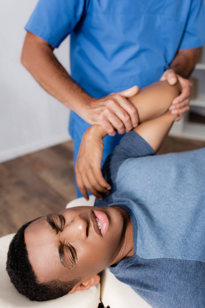 quiroprático trabalhando com braço de paciente afro-americano ferido na mesa de massagem - Foto, Imagem