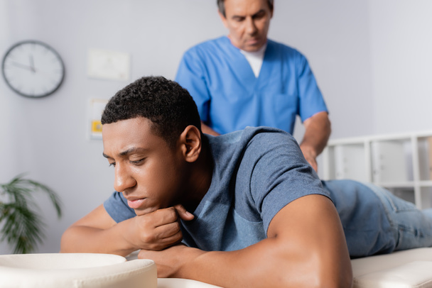 van middelbare leeftijd chiropractor werken met jong Afrikaans Amerikaans patiënt op massage tafel - Foto, afbeelding