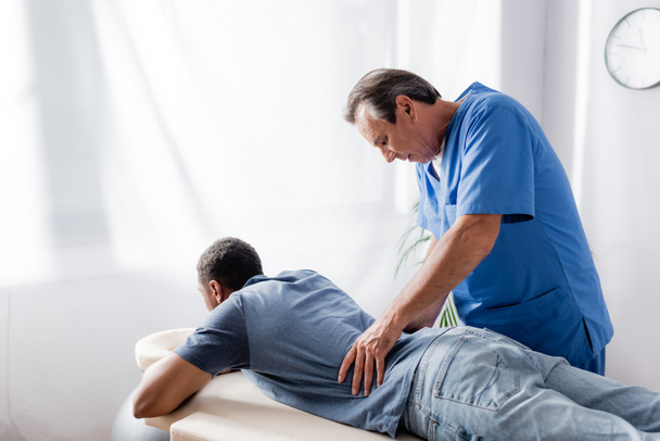 chiropratico di mezza età che lavora con un giovane e ferito paziente afroamericano sul lettino da massaggio in clinica - Foto, immagini