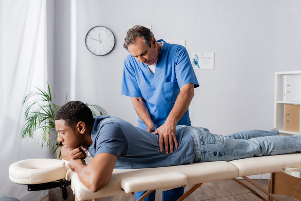 quiropráctico de mediana edad que trabaja con la espalda lesionada del paciente afroamericano en la mesa de masaje en la clínica - Foto, imagen