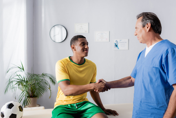 Glücklicher afrikanisch-amerikanischer Fußballer schüttelt Arzt in Klinik die Hand  - Foto, Bild