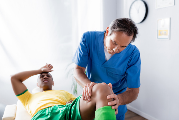 middle aged masseur massaging knee of african american sportsman on blurred background  - Photo, Image