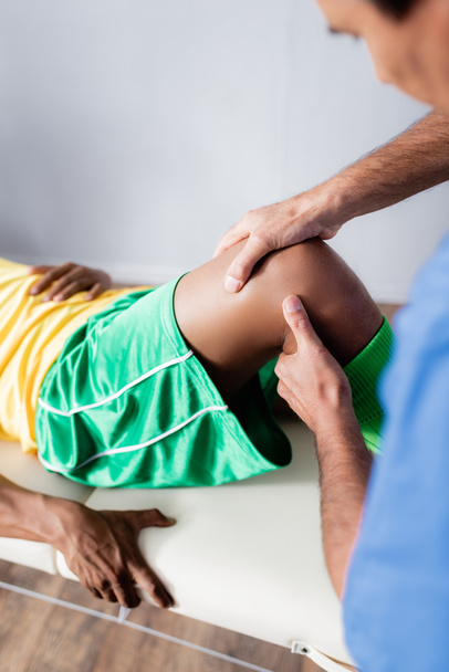 massagista de meia idade desfocado massageando joelho de esportista afro-americano em sportswear na clínica  - Foto, Imagem
