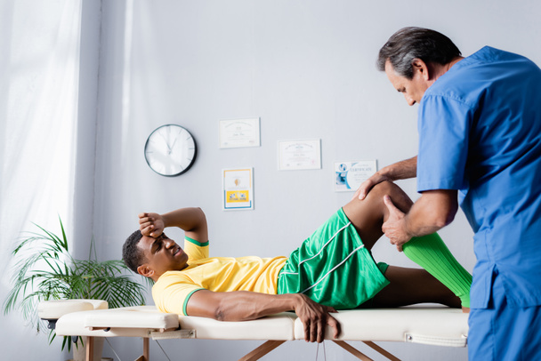 masajista de mediana edad masajeando rodilla de herido afroamericano deportista en ropa deportiva en clínica  - Foto, Imagen