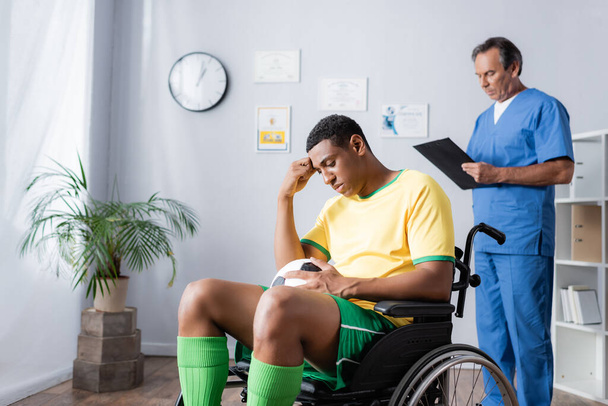 smutny afrykański amerykański sportowiec na wózku inwalidzkim trzymając piłkę nożną w pobliżu lekarza na rozmytym tle - Zdjęcie, obraz