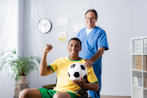 cheerful african american sportsman in wheelchair holding football and smiling with doctor  - Photo, image