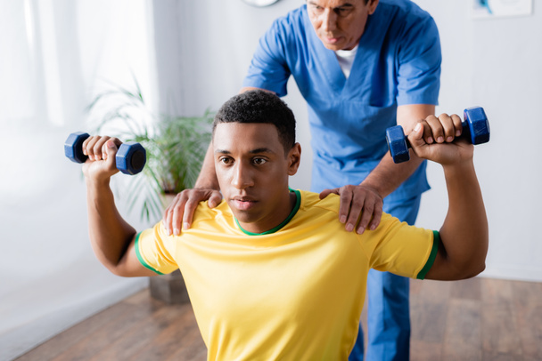 african american sportsman exercising with dumbbells near physiotherapist on blurred background  - Fotó, kép