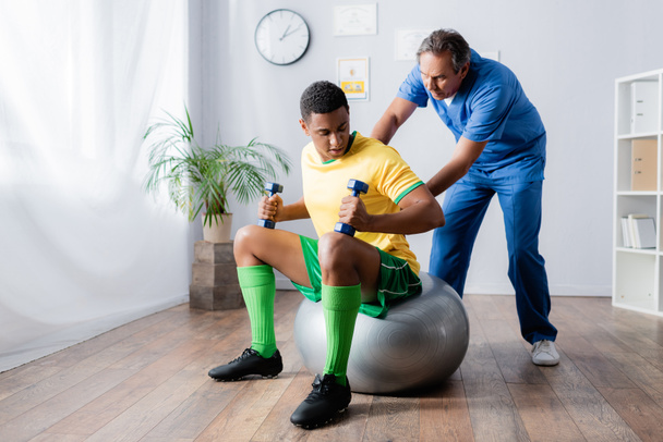 african american sportsman exercising with dumbbells on fitness ball near physiotherapist in clinic  - Photo, image