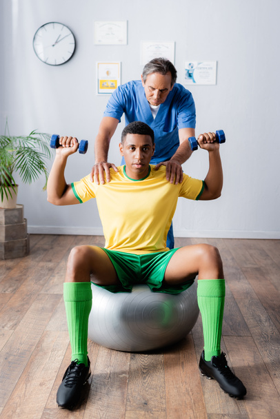 afro-amerikai sportoló edzés súlyzókkal fitnesz labdán fizioterapeuta közelében rehabilitáció alatt  - Fotó, kép