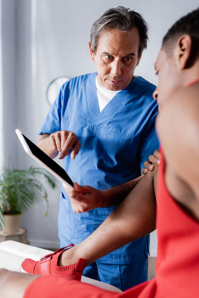 chiropractor pointing with finger at digital tablet near african american man in sportswear on blurred foreground - Foto, imagen