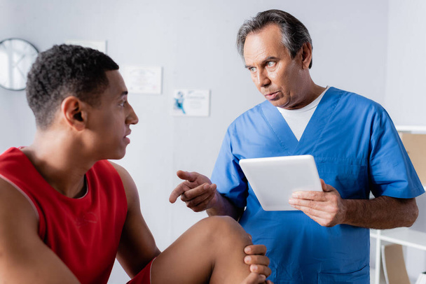 chiropracteur tenant tablette numérique tout en parlant et en gestuant près d'un patient afro-américain en vêtements de sport  - Photo, image