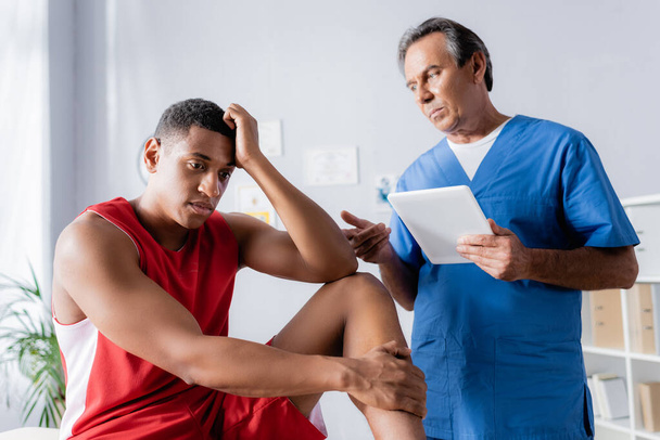 χειροπρακτικός κρατώντας ψηφιακή ταμπλέτα κοντά αναστατωμένος Αφροαμερικανός άνδρας σε αθλητικά είδη στην κλινική - Φωτογραφία, εικόνα