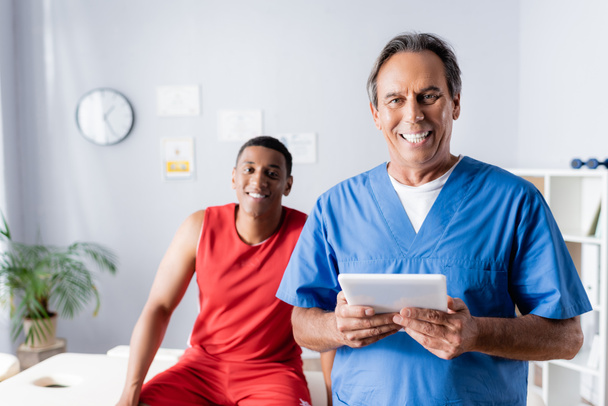 alegre médico sosteniendo tableta digital cerca feliz africano americano paciente en ropa deportiva sobre fondo borroso  - Foto, Imagen