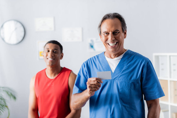 happy doctor holding blank card near african american patient in sportswear on blurred background  - 写真・画像