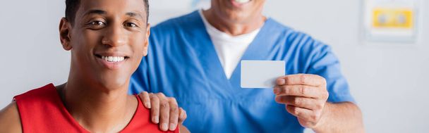 doctor borroso sosteniendo la tarjeta en blanco y tocando el hombro del paciente afroamericano feliz, bandera - Foto, Imagen