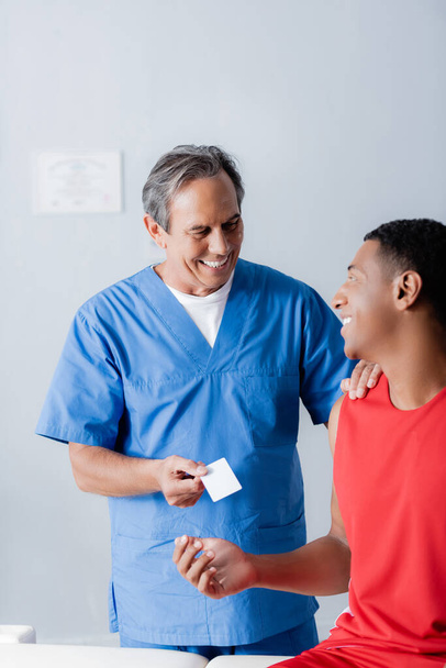 feliz doctor dando en blanco tarjeta a alegre afroamericano deportista  - Foto, Imagen