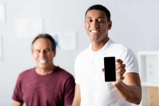 веселый африканский американский терапевт держит смартфон с пустым экраном рядом с пациентом на размытом фоне  - Фото, изображение