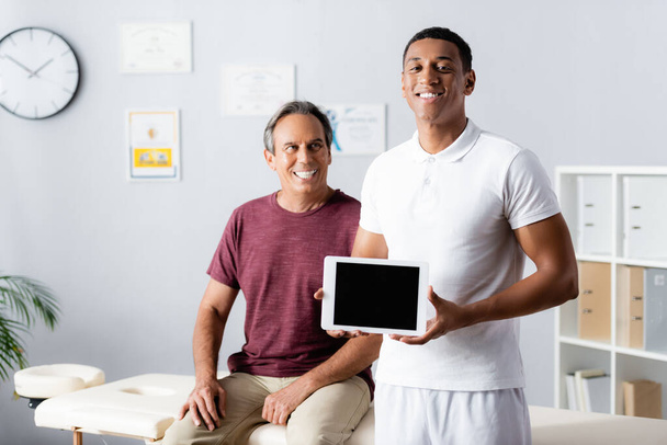 vrolijk Afrikaans amerikaanse therapeut houden digitale tablet met blanco scherm in de buurt patiënt op wazige achtergrond  - Foto, afbeelding