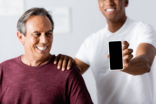 ευτυχισμένη Αφροαμερικανός θεραπευτής κρατώντας smartphone με λευκή οθόνη κοντά στον ασθενή σε θολή φόντο  - Φωτογραφία, εικόνα