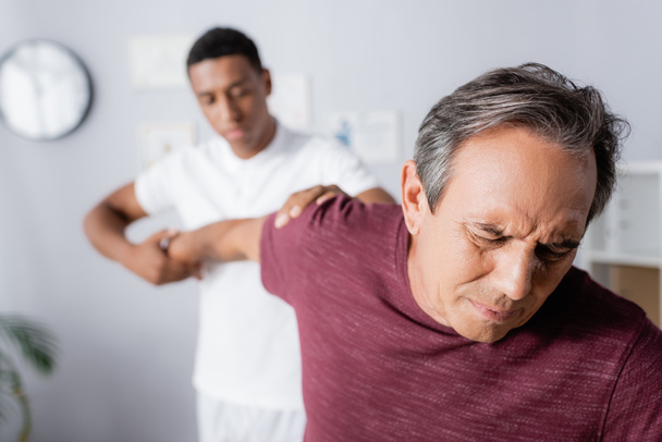 homem maduro que sofre de dor ao receber massagem do fisioterapeuta afro-americano em fundo turvo - Foto, Imagem