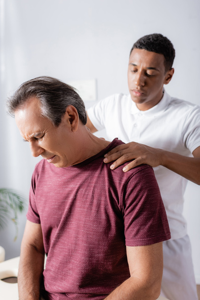 fisioterapista afroamericano che massaggia l'uomo di mezza età con gli occhi chiusi in clinica - Foto, immagini