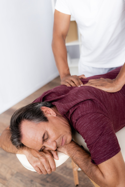 young african american masseur massaging back of middle aged man on massage table - 写真・画像