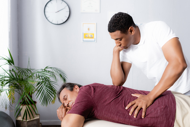 african american physiotherapist massaging back of middle aged man on massage table - Foto, afbeelding