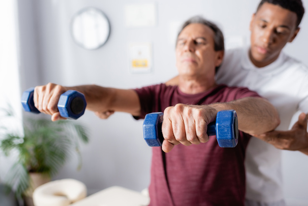 hombre de mediana edad haciendo ejercicio con pesas cerca de fisioterapeuta afroamericano sobre fondo borroso  - Foto, Imagen
