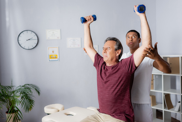középkorú férfi edz súlyzók közelében afro-amerikai fizioterapeuta  - Fotó, kép