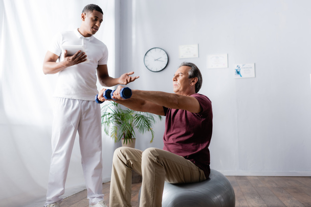 middle aged man exercising with dumbbells on fitness ball near african american physiotherapist with digital tablet  - Valokuva, kuva