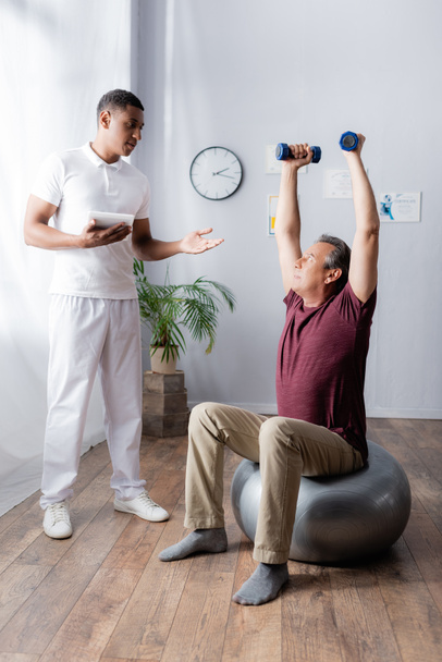 middle aged man exercising with dumbbells on fitness ball near african american physiotherapist with digital tablet pointing with hand in clinic - Photo, image