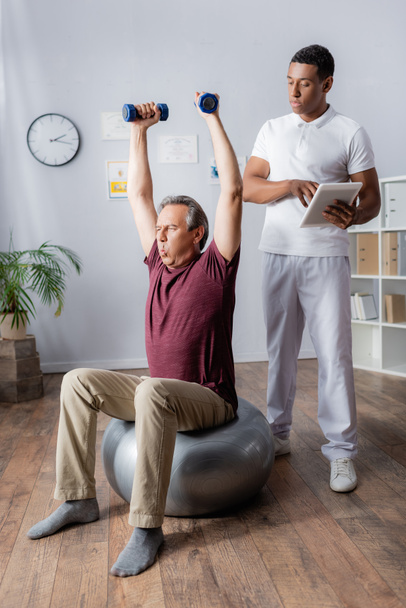 african american physiotherapist with digital tablet looking at middle aged man exercising with dumbbells on fitness ball  - Φωτογραφία, εικόνα